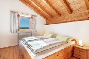 Un dormitorio con techo de madera, ventana con cortinas, cama con almohadas, mesita de noche con lámpara y radiador.
