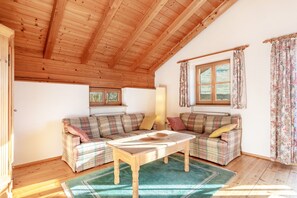 A cozy room with wooden ceilings, plaid sofas, a wooden coffee table, and a large window with curtains.
