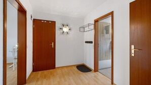Un pasillo con suelo de madera, una puerta con interruptor de luz, una lámpara fijada a la pared y un felpudo en la entrada.