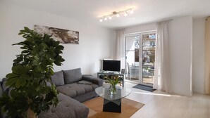 A modern living room with a grey sofa, a glass coffee table, a large plant, a flat-screen TV, and a sliding glass door leading to an outdoor area. - Quiet Apartment in Bad Saulgau with Mountain View, Private Terrace and Wi-Fi (Bad Saulgau)
