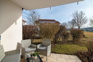 Une terrasse dotée de meubles en osier, d’une table ornée d’une plante, et offrant une vue sur une maison et des arbres.