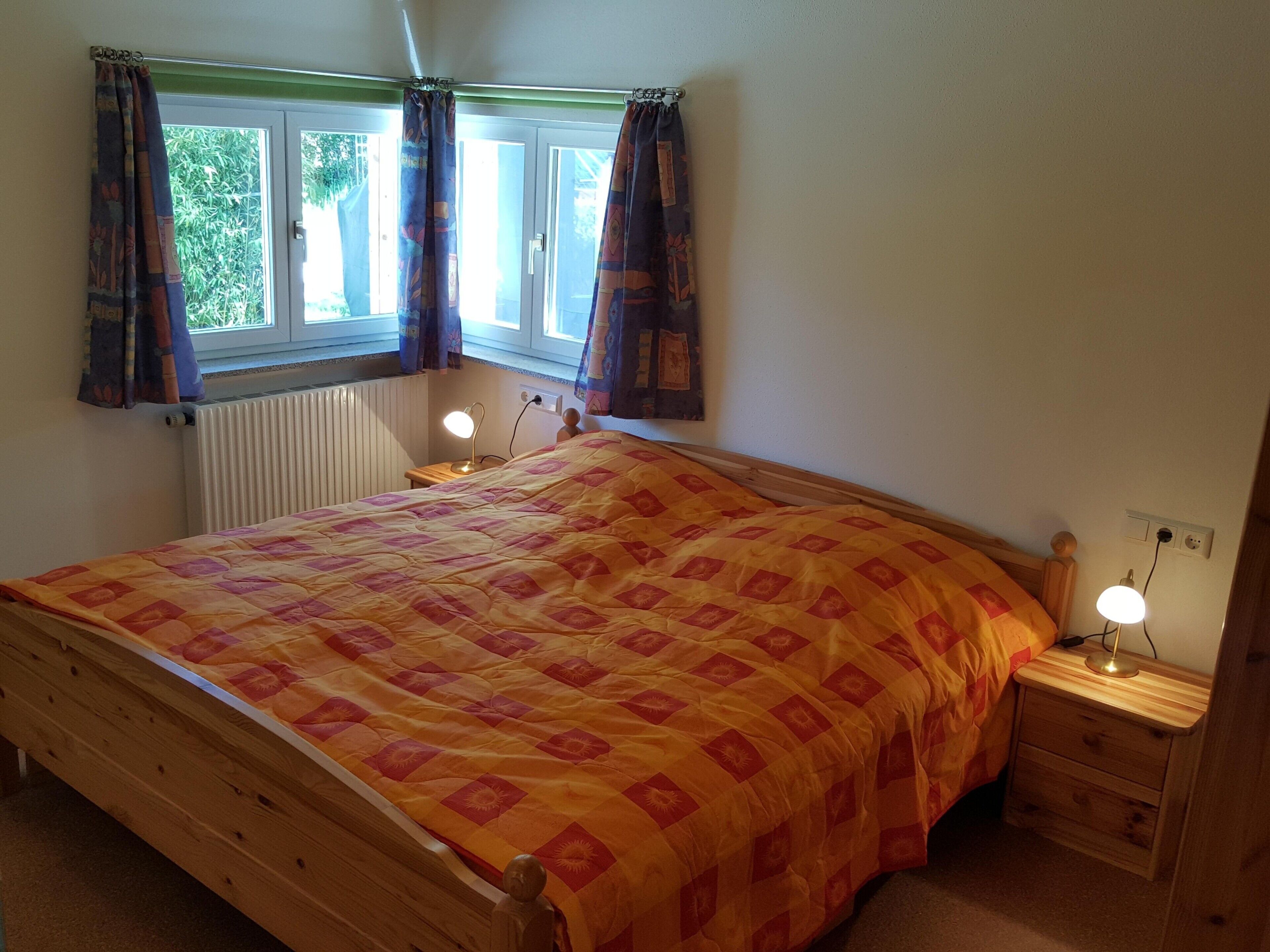 A bedroom with a wooden bed, two bedside lamps, a window with curtains, and a radiator.
