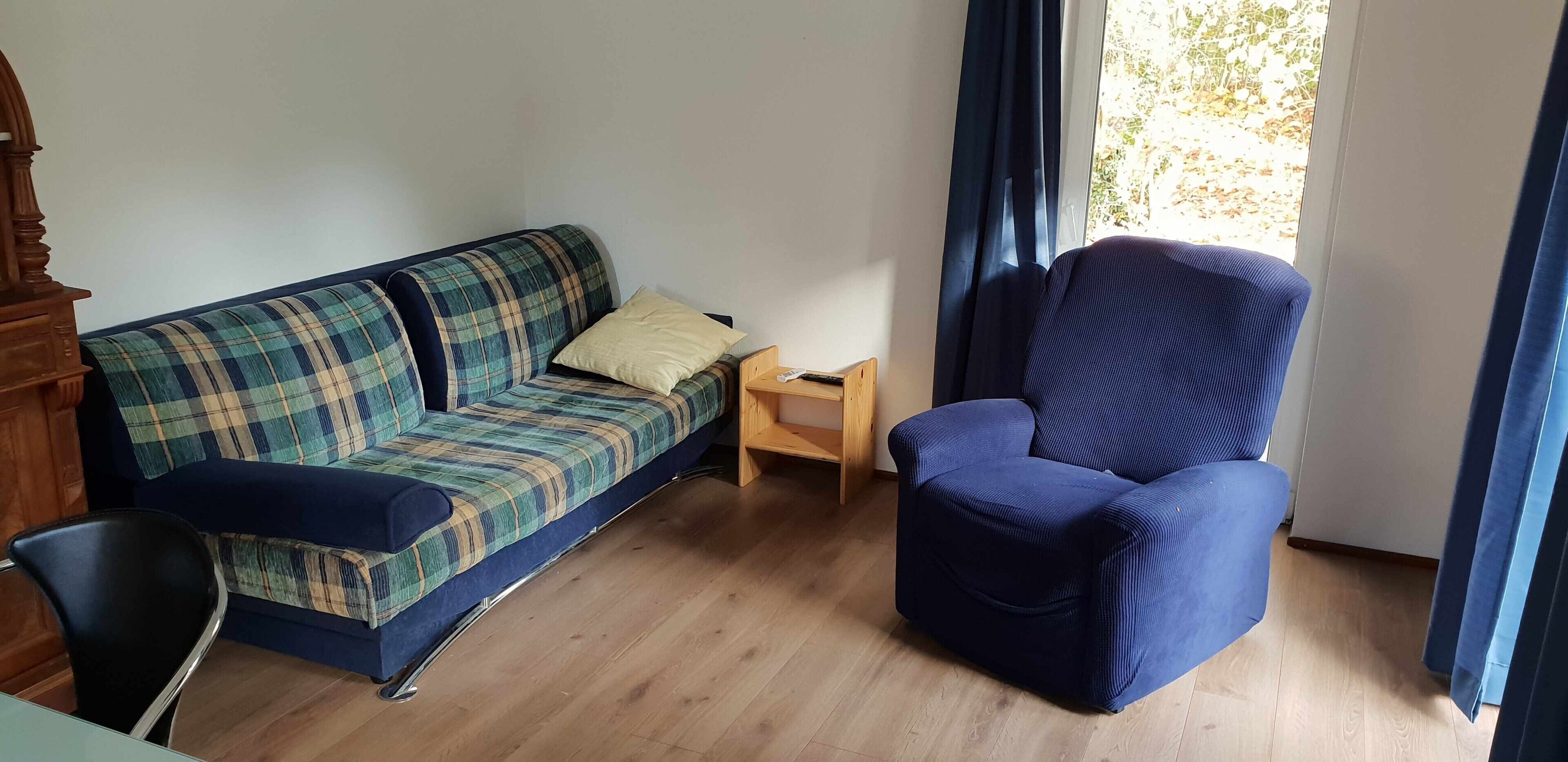 Una habitación con un sofá a cuadros, un sillón azul, una mesita de noche de madera y una ventana con cortinas.