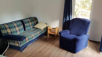 Una habitación con un sofá a cuadros, un sillón azul, una mesita de noche de madera y una ventana con cortinas.