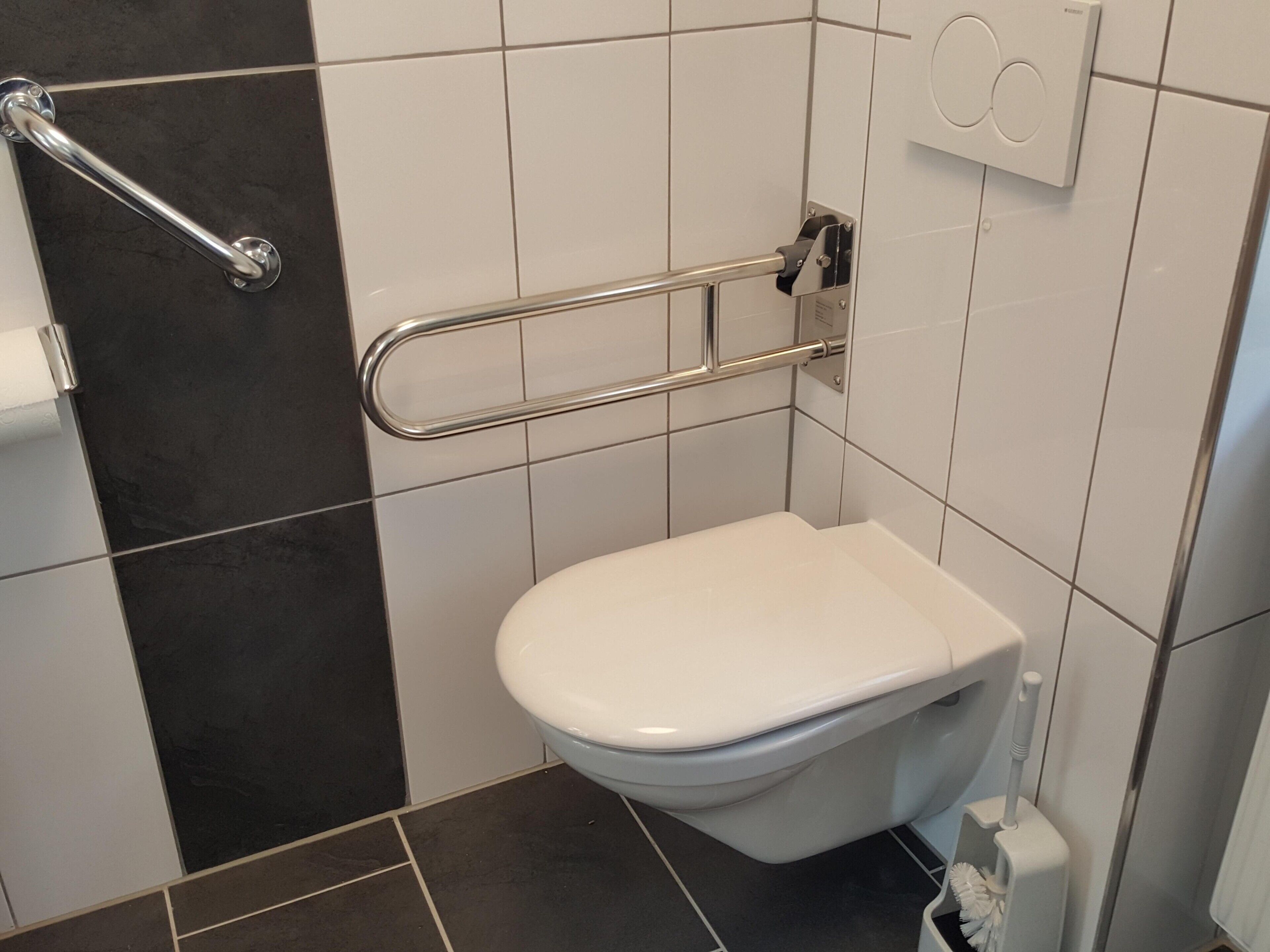A bathroom with a white toilet, a handrail, a black-tiled wall, and a white floor.