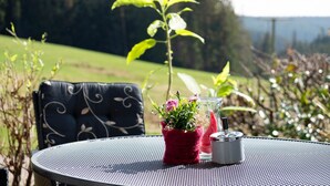 Una mesa metálica redonda con una silla tejida, una maceta roja con flores y un recipiente de vidrio con tapa de metal.