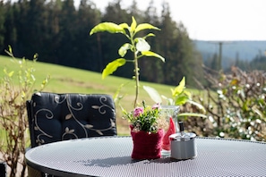A round metal table with a woven chair, a red pot with flowers, and a glass container with a metal lid.