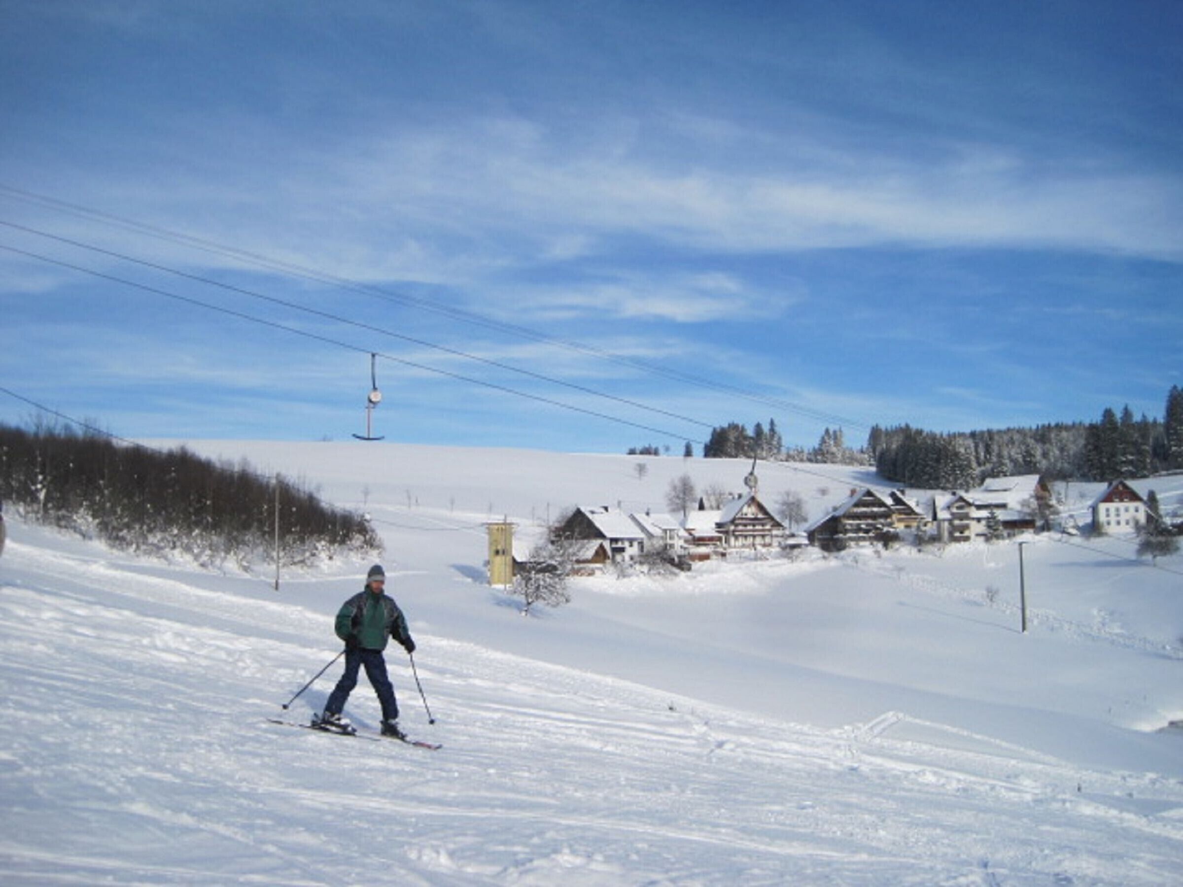 A snowy ski slope with a person skiing, houses in the background, and a ski lift.