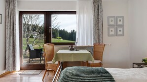 Una habitación con una cama, una mesa con un florero, sillas de mimbre y vistas a un jardín a través de un ventanal grande.