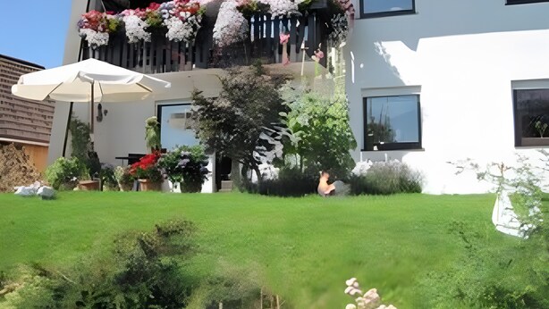 Un edificio de dos pisos con un balcón decorado con flores, una sombrilla blanca y un césped bien cuidado.