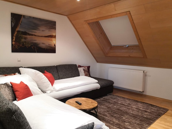 A loft-style room with a sloped ceiling, a large window, a grey sofa, a small round table, and a radiator.