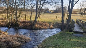A rural stream with a wooden bridge, surrounded by trees and a grassy field.