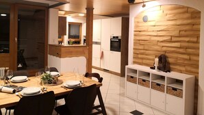 A modern kitchen with wooden cabinetry, a dining area with a wooden table set for four, and a built-in white shelving unit.