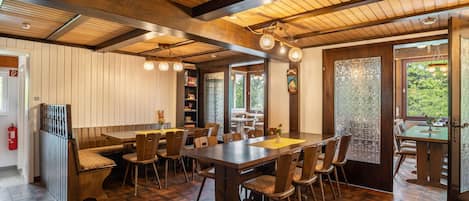 A dining area with wooden tables and chairs, a high ceiling with exposed beams, and large windows with a view of greenery.