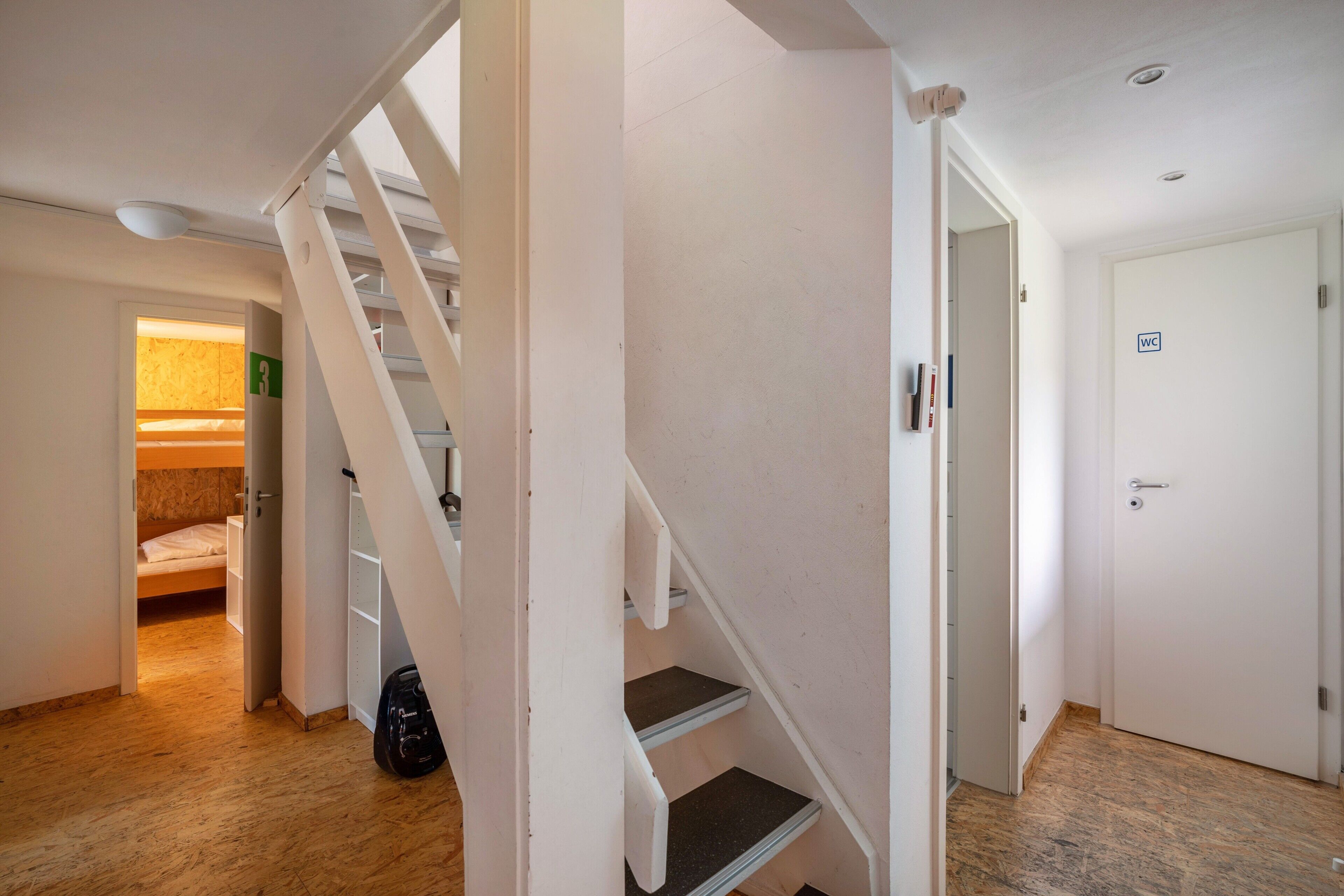 A staircase leading to a bedroom with a bunk bed, a door with a WC sign, and a bag on the floor.