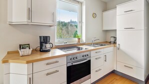 Eine moderne Küche mit weißen Schränken, einem Backofen, einer Kaffeemaschine und einem Fenster mit Blick auf Bäume.