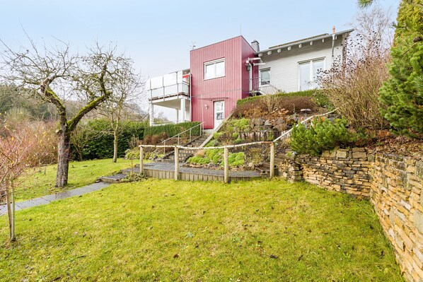 Uma casa moderna com fachada vermelha, um muro de pedra e um jardim gramado com uma escadaria de madeira que leva à entrada.
