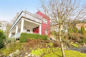 Um edifício moderno de dois andares com varanda, fachada vermelha e um caminho de pedra que leva à entrada.