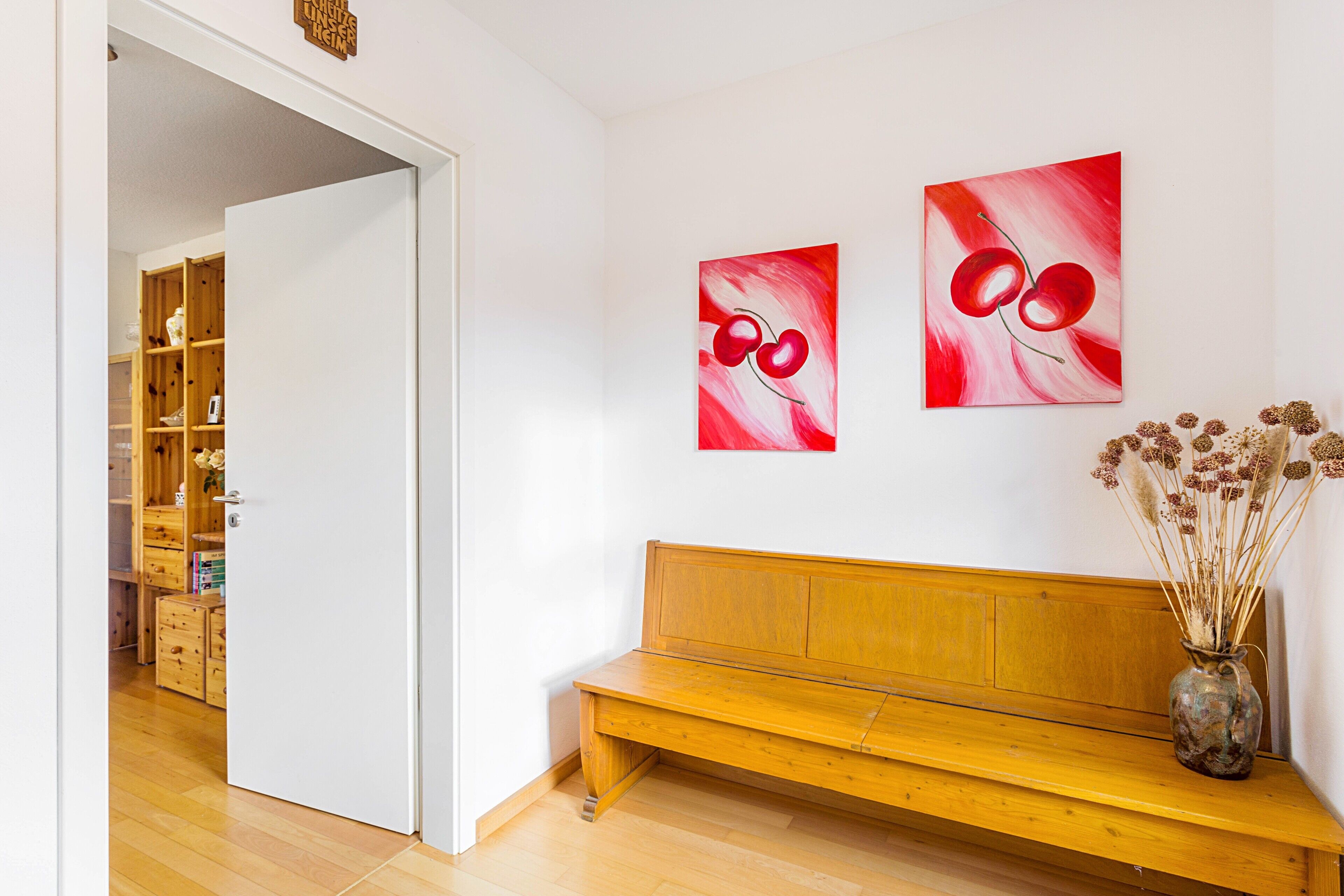 Ein Zimmer mit einer Holzbank, einer Vase mit Trockenblumen und zwei roten abstrakten Bildern an der Wand.