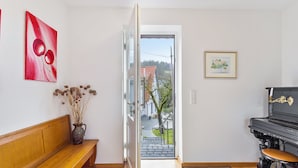 Ein Zimmer mit einer hölzernen Bank, einer Vase mit Trockenblumen, einem roten abstrakten Gemälde, einem Klavier und einer Tür zu einem Außenbereich mit Baum und Haus.