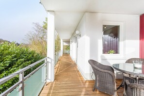 Uma área externa coberta com deck de madeira, móveis de vime e uma mesa de vidro.