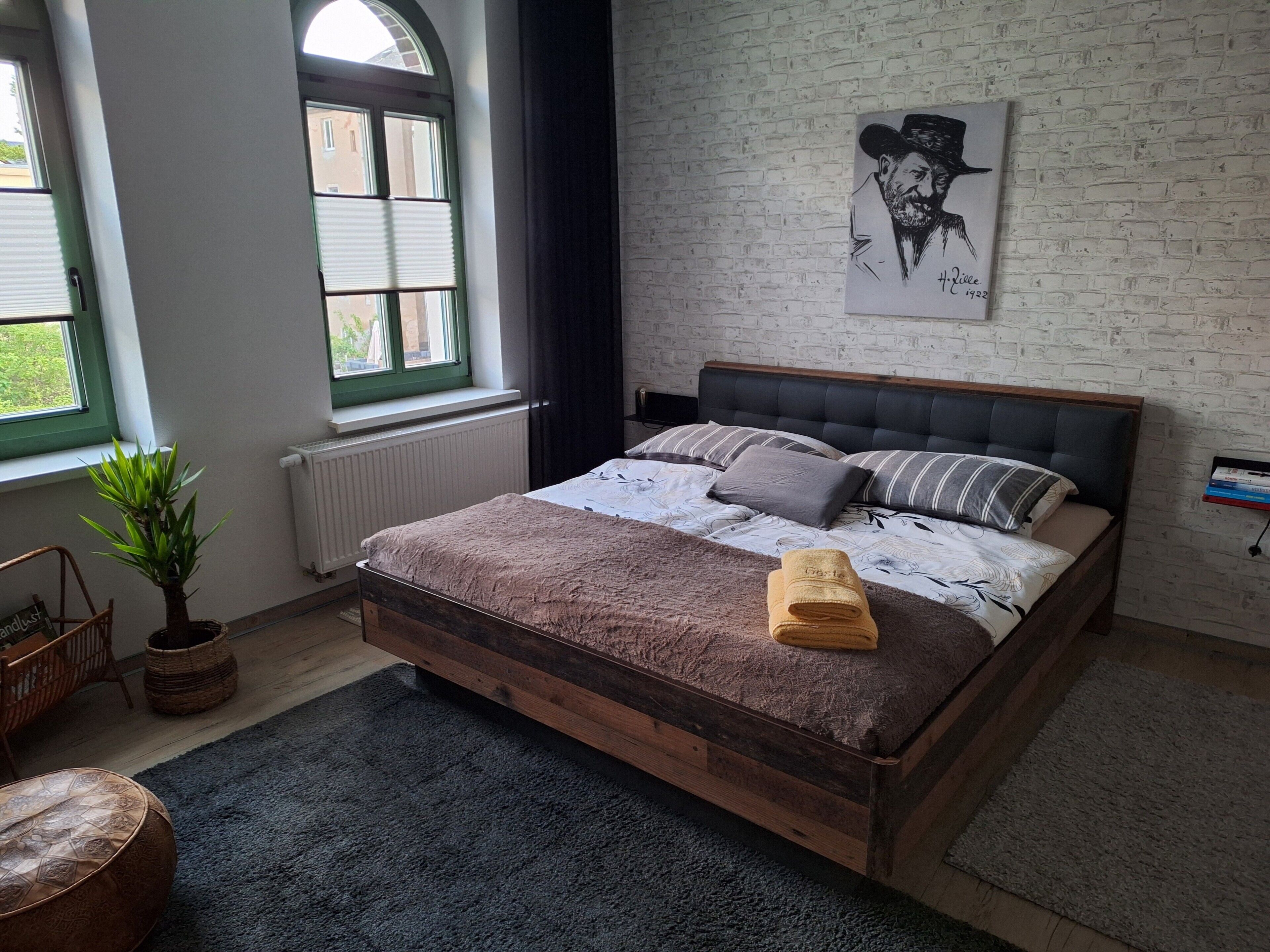 A bedroom with a wooden bed, a window with blinds, a potted plant, a wall-mounted artwork, and a bedside table.