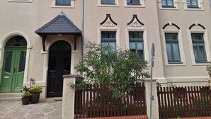 Ein Gebäude mit einer symmetrischen Fassade, zahlreichen Fenstern mit Fensterläden, einem zentralen Eingang mit einem dekorativen Bogen und einer kleinen Gartenanlage mit Pflanzgefäßen.