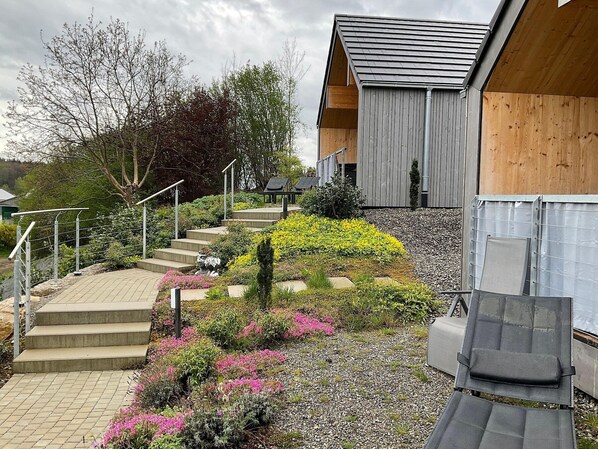 Un edificio moderno con fachada de madera, una escalera de hormigón que conduce a una terraza de madera, y un jardín con diversas plantas y flores.