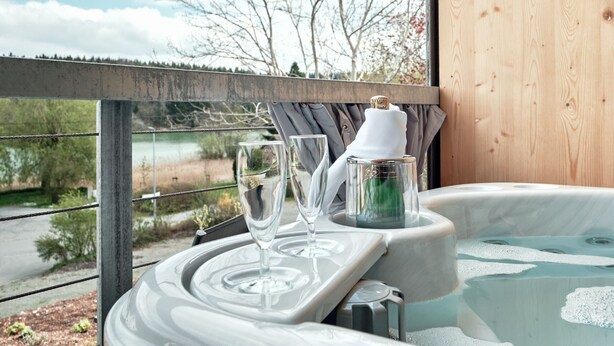Un jacuzzi con vistas a un lago y árboles, una terraza de madera y una mesa con vasos y una botella.