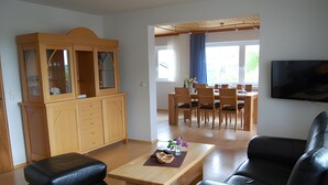 A living room with a wooden cabinet, a black leather sofa, a wooden coffee table, and a dining area with wooden chairs.