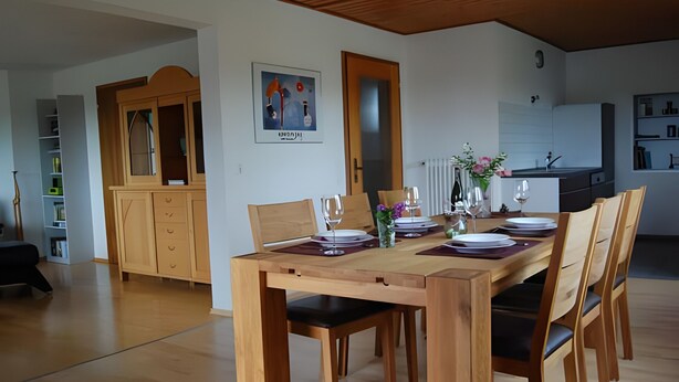 A dining area with a wooden table set for four, a cabinet with glass doors, and a kitchen in the background.