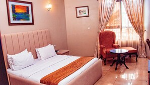 A hotel room with a bed, a chair, a small table, and two framed pictures on the wall. - DEES HOTELS APARTMENT & SUITES (Lagos)