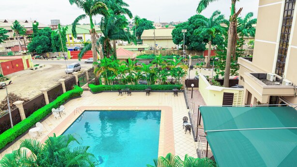 A swimming pool surrounded by lush greenery and palm trees, with a view of the surrounding buildings. - DEES HOTELS APARTMENT & SUITES (Lagos)