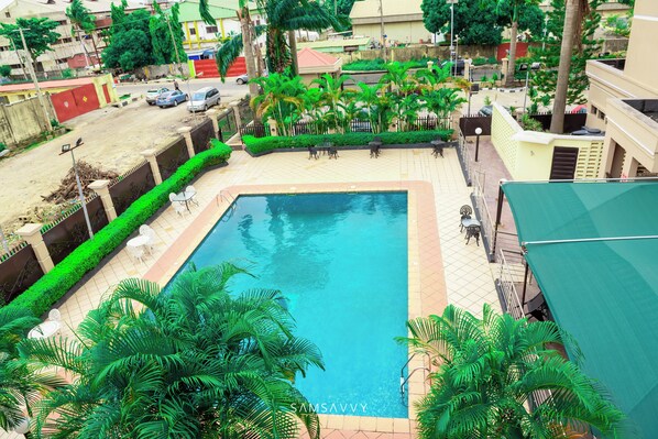A rectangular swimming pool with a tiled border, surrounded by a low wall and lush green palm trees. - DEES HOTELS APARTMENT & SUITES (Lagos)