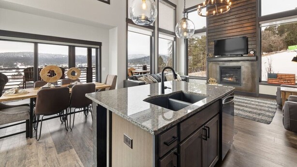 A modern kitchen with a large island, dining area with chairs, and a fireplace. - Modern Mountain Retreat (Sturgis)