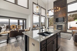 A modern kitchen with a large island, dining area with chairs, and a fireplace. - Modern Mountain Retreat (Sturgis)