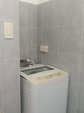 A compact, white washing machine in a corner with grey tiled walls, a visible electrical outlet, and a water inlet pipe. - Spacious 2BR at Sea Residences Near MOA - Family Stay (Pasay)
