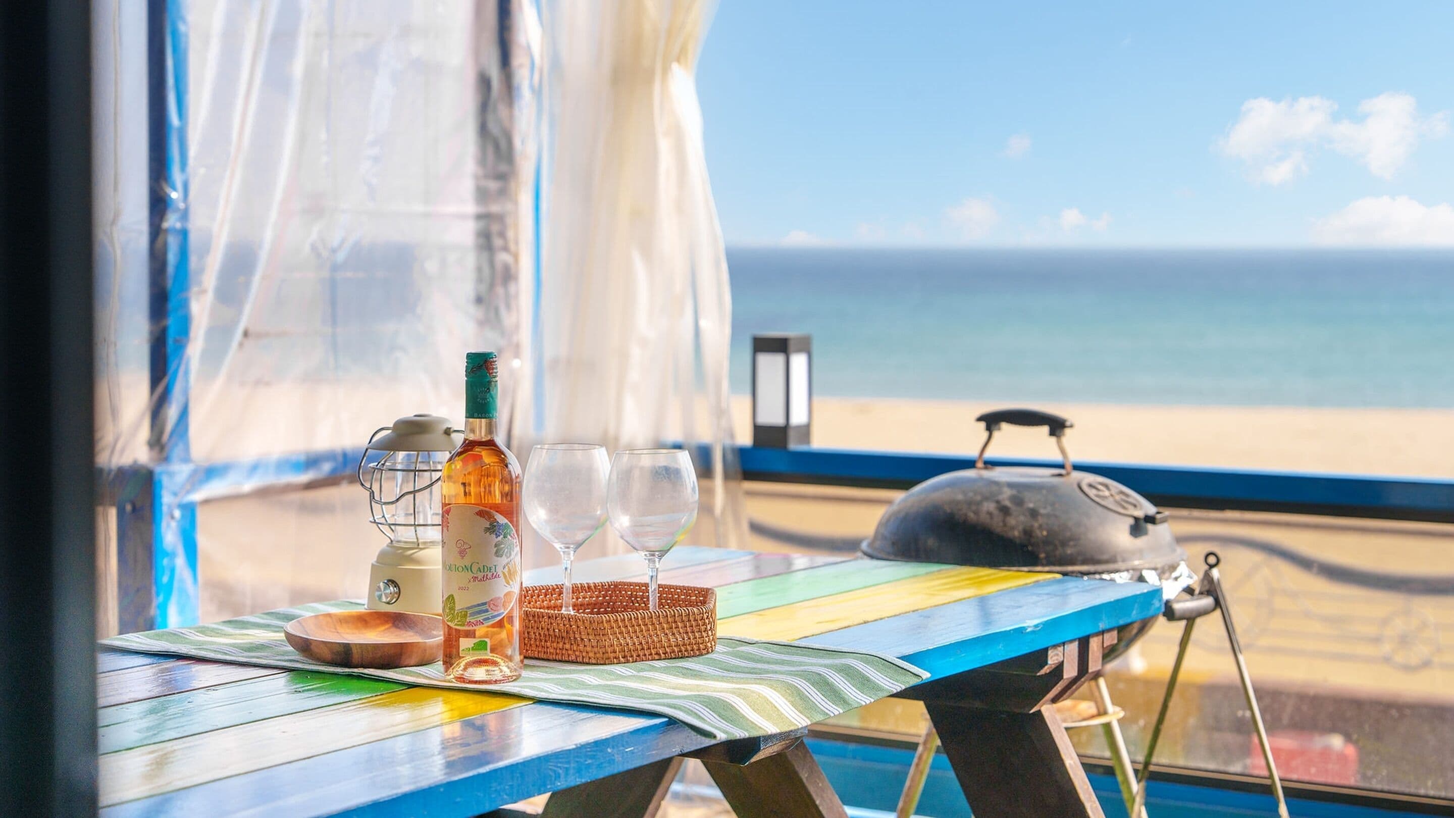 Uma mesa com uma garrafa, copos, um cesto e uma chaleira, tudo próximo a uma janela com vista para a praia.