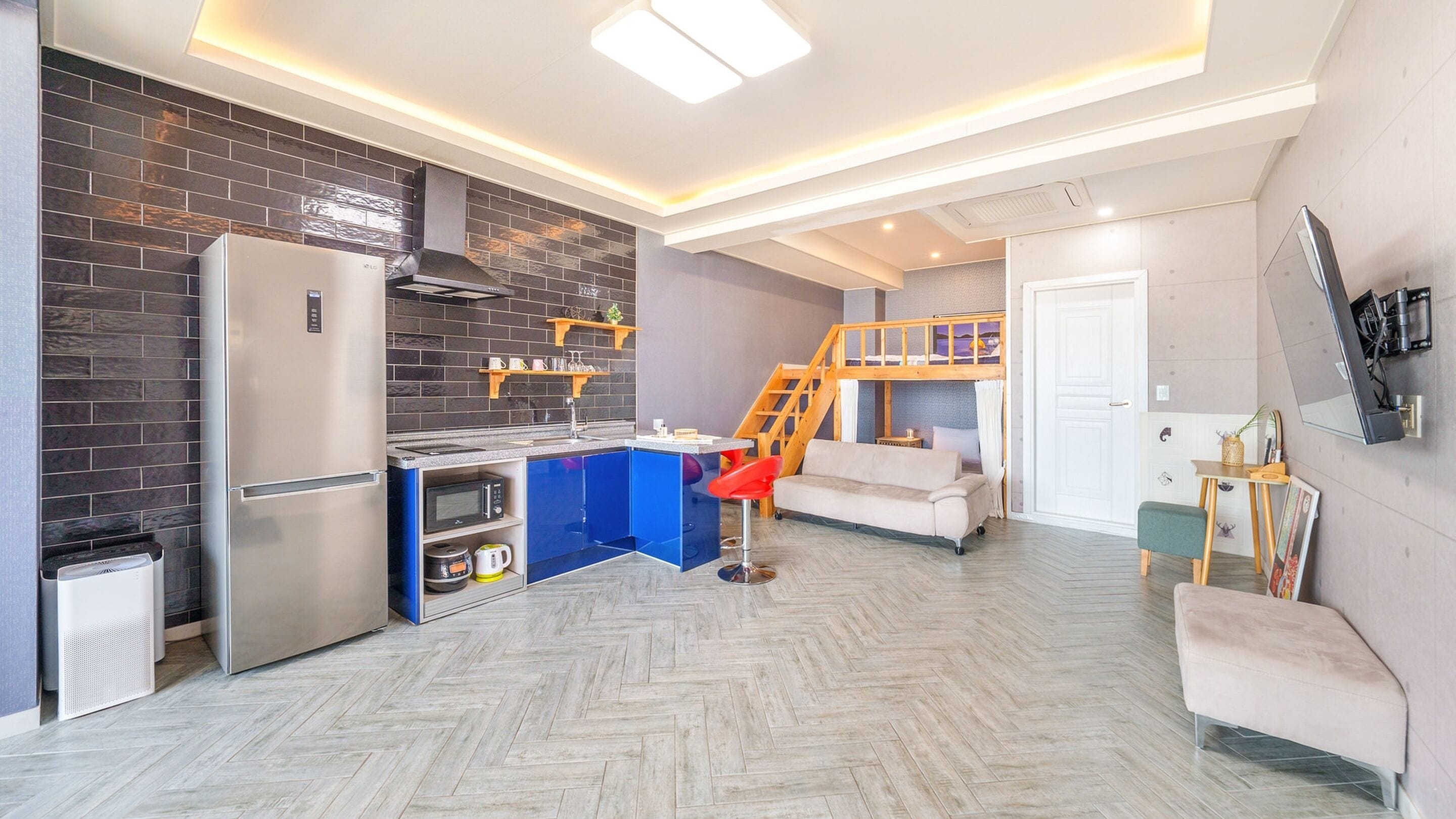 Un espacio habitable pequeño con kitchenette, un sofá, una silla roja, una escalera de madera y una puerta.
