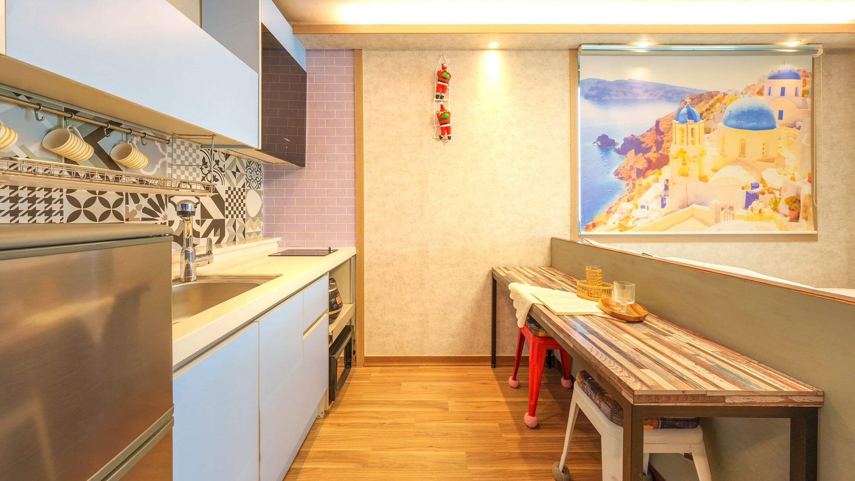 A compact kitchen with a modern design, featuring a stainless steel refrigerator, a sink, and a patterned backsplash. A wooden table with a tray and glass is positioned near a window displaying a coastal scene.