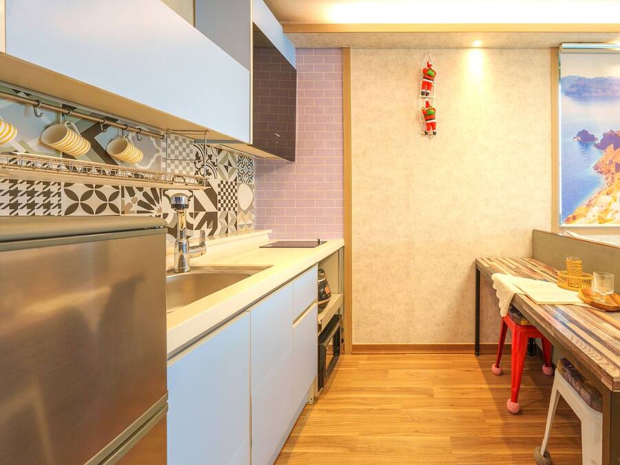 A compact kitchen with a modern design, featuring a stainless steel refrigerator, a sink, and a patterned backsplash. A wooden table with a tray and glass is positioned near a window displaying a coastal scene.