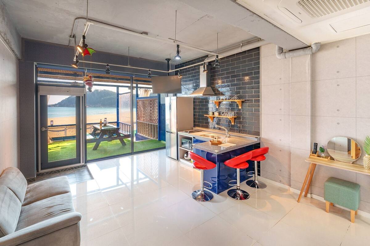 Une cuisine moderne dotée d’un grand îlot central, de tabourets de bar rouges et offrant une vue sur la plage grâce à de grandes fenêtres.