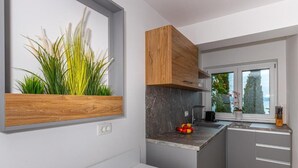 A modern kitchen with grey cabinets, a wooden upper cabinet, a marble countertop, and a window with a view of trees. - Apartman Helena (Crikvenica)