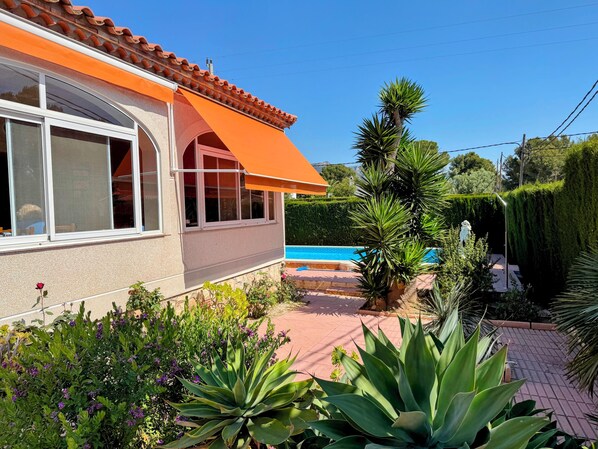 A house with a swimming pool, surrounded by various plants and a clear blue sky.