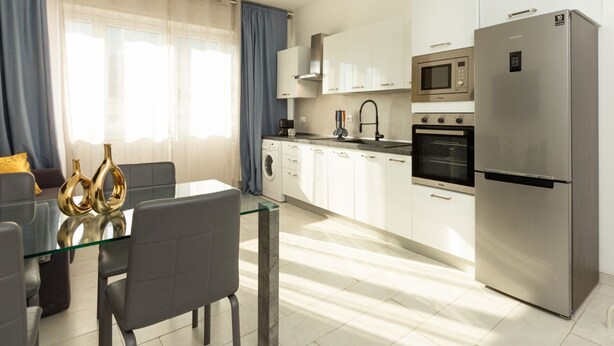 Een moderne keuken met witte kastjes, een roestvrijstalen koelkast, een magnetron en een oven. Er staat een glazen eettafel met stoelen en decoratieve vazen.