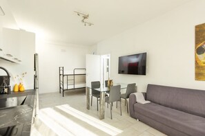Una sala de estar moderna con un sofá gris, un televisor de pantalla plana montado en la pared y un comedor con una mesa y sillas de vidrio.