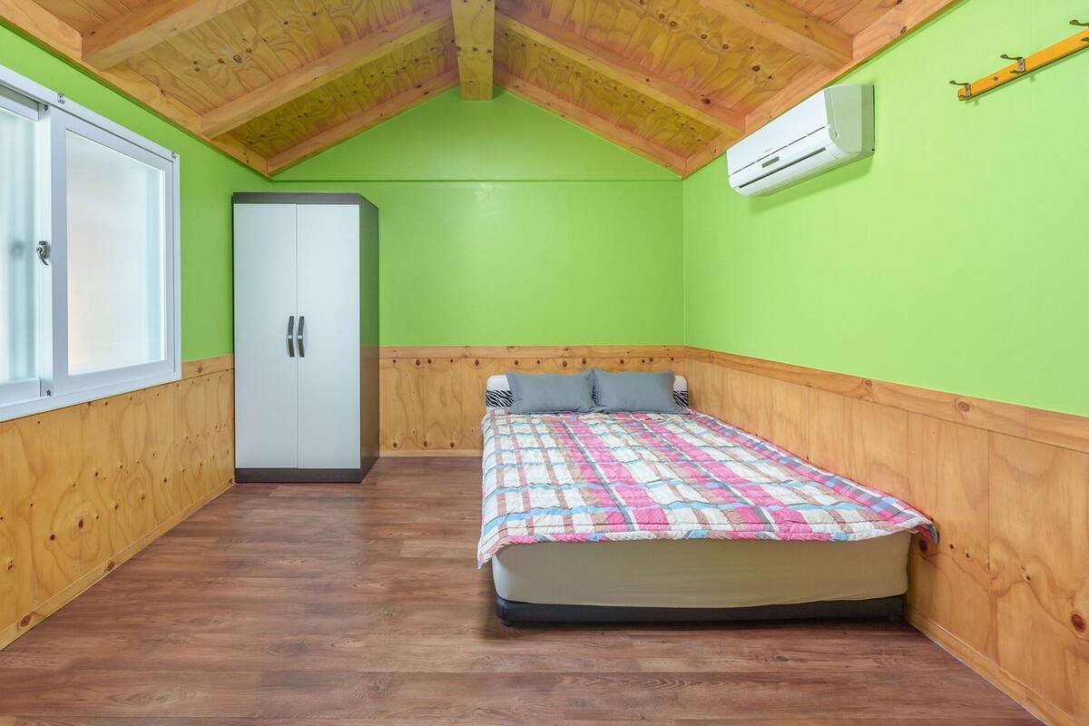 A bedroom with a wooden ceiling, a bed with a colorful quilt, a wardrobe, and a window.