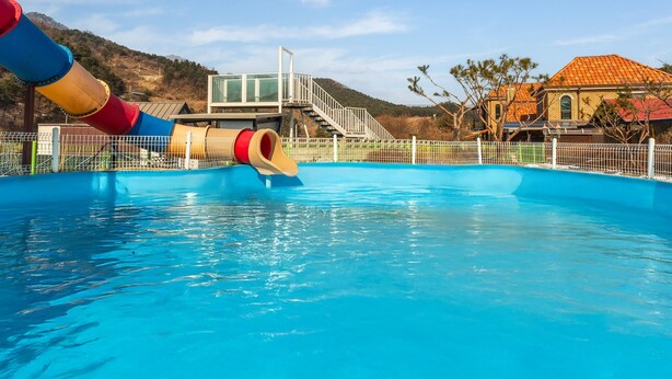 A swimming pool with a water slide, surrounded by a white fence, and a building with a red roof in the background. - Gapyeong Sanneomeo Private Pension (Gapyeong)