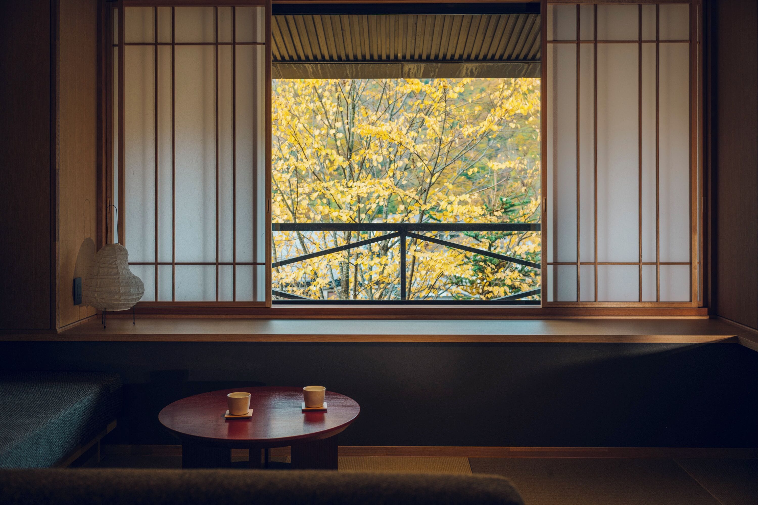A traditional Japanese room with tatami flooring, a low table with a cup, and a view of autumn foliage through shoji screens.