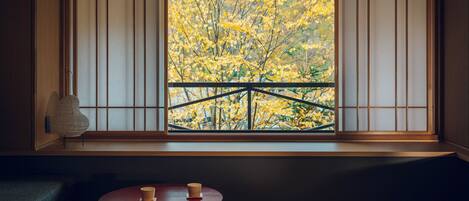 Una habitación japonesa tradicional con suelo de tatami, una mesa baja con una taza y vistas a follaje otoñal a través de celosías shoji.